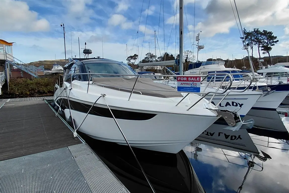 A sleek white boat docked at a marina with a "FOR SALE" sign, surrounded by other vessels under a cloudy sky.